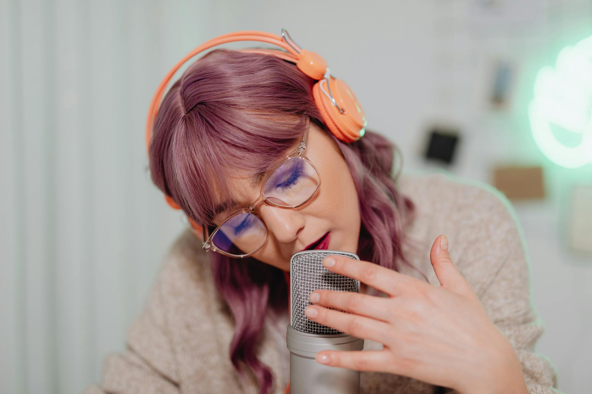 A young woman with vibrant hair and glasses recording audio with a microphone indoors.